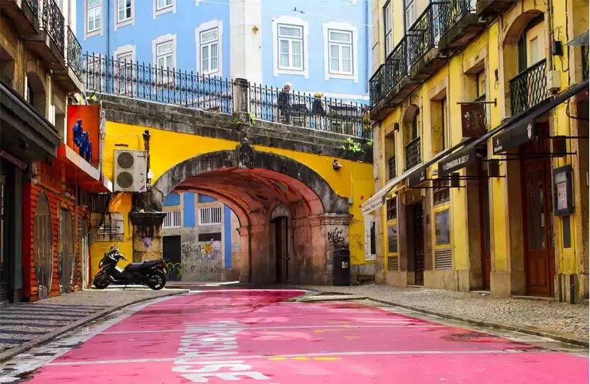 Em dia de sol, moto estacionada em rua com asfalto rosa e construções coloridas em volta