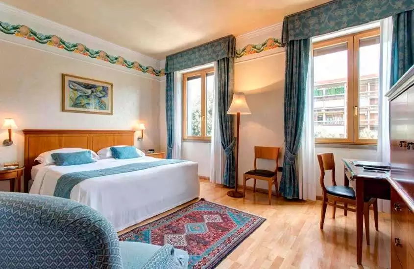 Durante o dia, quarto de um dos hotéis em verona com cama de casal, janelas grandes acortinadas, decoração em tons de madeira e azul, quadro, luminárias, cadeiras e área de trabalho