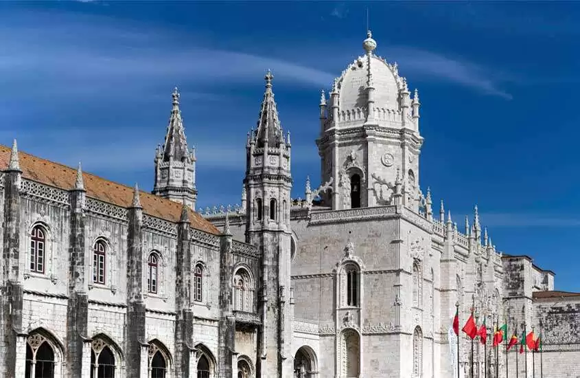 Em final de tarde, vista da lateral do Mosteiro dos Jerônimos, ponto turístico em Lisboa do século XV, e bandeiras de Portugal hasteadas a direita