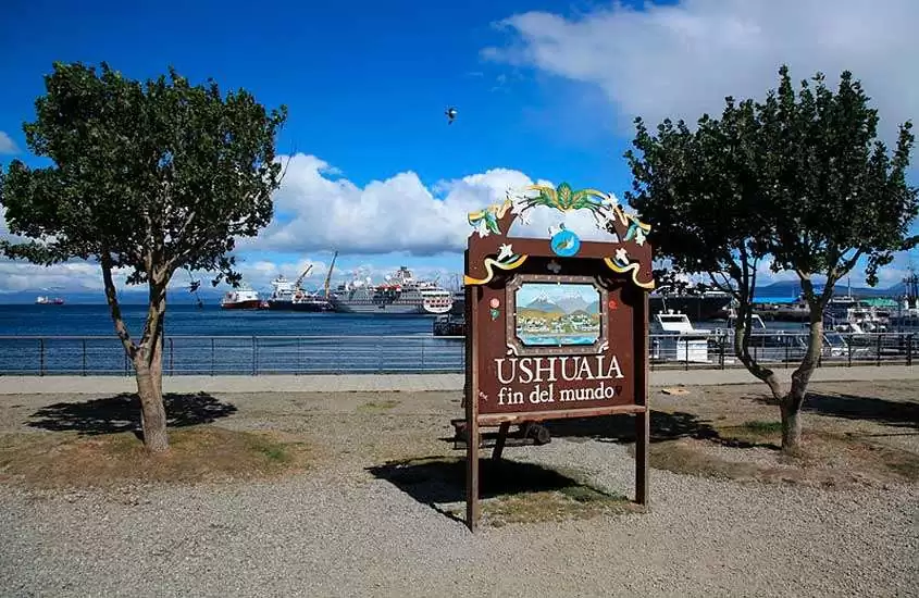 Durante o dia, placa de madeira escrita ''Ushuaia fin del mundo'', um dos pontos turísticos de Ushuaia. Ao fundo, mar com barcos