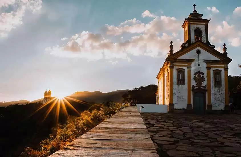 em dia ensolarado, igreja barroca a direita e montanhas ao fundo.