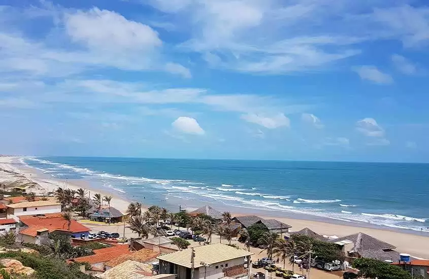 durante dia de sol, vista panorâmica da orla de fortaleza com cabanas, casas, palmeiras e mar azul.