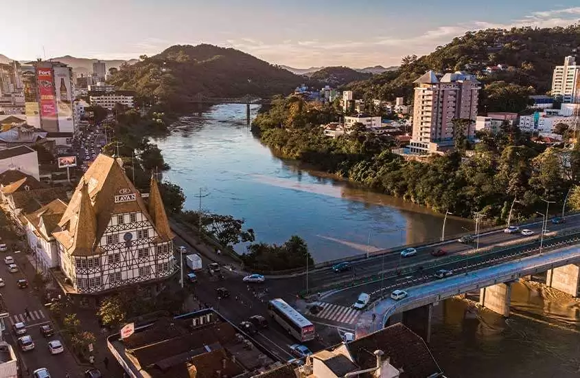 em final de tarde, vista panorâmica aérea de Blumenau com canal no meio, árvores, serra no fundo e construções ao redor