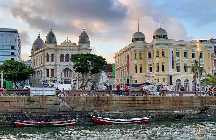 durante o dia, pessoas sentadas em mureta às margens de canal em Recife.