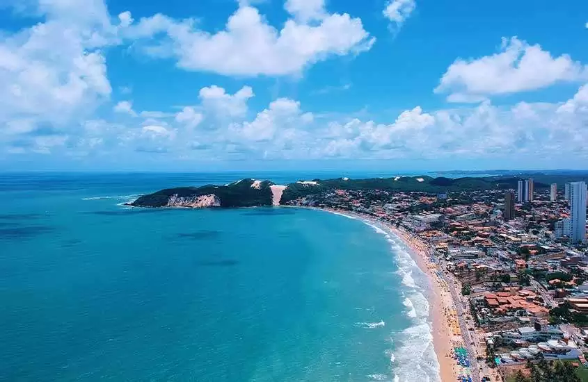 em dia de sol com nuvens, vista aérea panorâmica de casas e prédios em frente à orla de natal com mar azul e ampla faixa de areia.