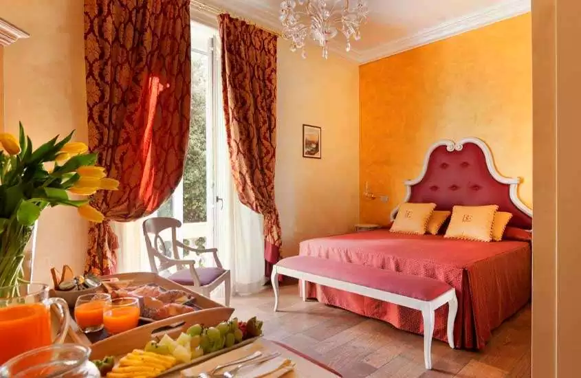 Durante o dia, quarto em tons de laranja e vermelho com cama de casal, almofadas laranjas, mesa com bandejas de café da manhã, janela grande acortinada, lustre e vaso de flores