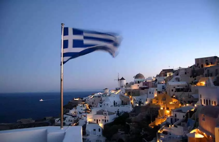 Ao anoitecer, bandeira da Grécia hasteada em muro. Ao fundo, casas às margens do mar