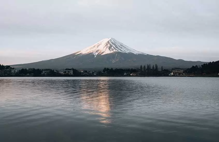 Durante um final de tarde, vista panorâmica de casas às margens de lago. Ao fundo, Monte Fuji, um dos pontos turísticos do japão, coberto de neve