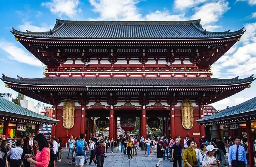 Em dia de sol, pessoas caminhando em frente a templo de Senso-Ji vermelho com detalhes em dourado e teto preto