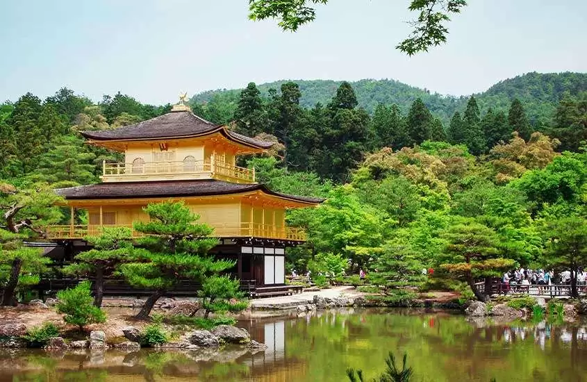 Em dia de clima ameno, templo de Pavilhão Dourado com lago em frente, árvores ao redor e montanhas ao fundo