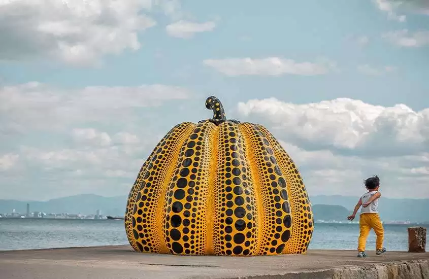 Em dia de sol, escultura de abóbora amarela com pontinhos pretos, mar e criança ao lado direito, em píer às margens do mar