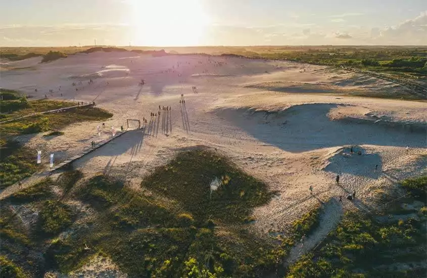 Em dia de sol, vista aérea de pessoas andando em dunas com área gramada ao redor