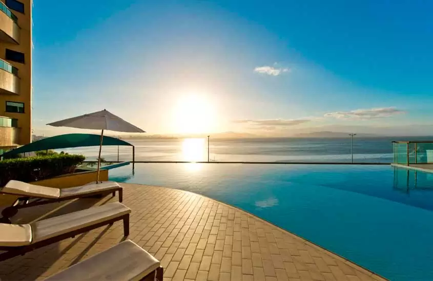 Em final de tarde de sol, espreguiçadeiras, guarda-sol e plantas em frente a piscina com vista para o mar em hotel para passar o réveillon florianópolis 2024