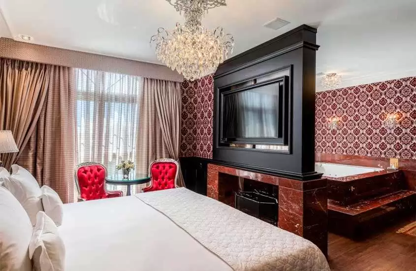 Quarto de um dos hotéis de luxo em gramado com cama de casal, mesa com cadeiras vermelhas, lareira, janela grande, lustre ornamentado e televisão. Ao fundo, banheira.
