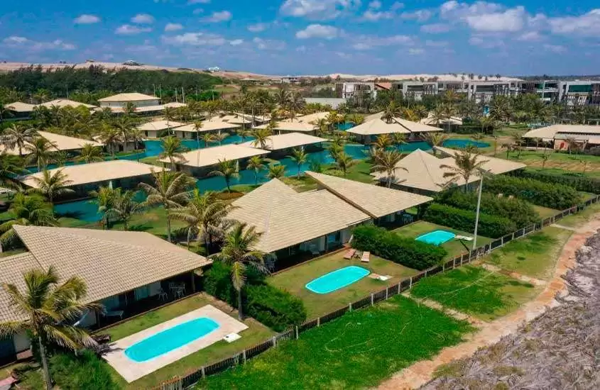 Em dia de sol, vista aérea de hotel com piscinas, chalés, área gramada e árvores ao redor