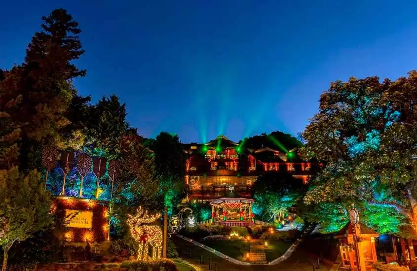 Durante a noite, vista aérea de um dos hotéis de luxo em gramado com lasers verdes, carrossel na frente, iluminação de natal e árvores ao redor