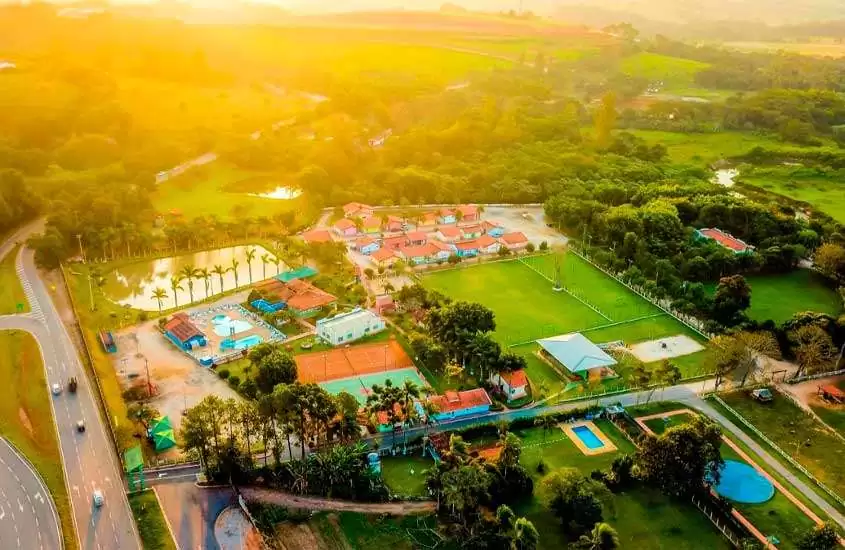 Em dia ensolarado vista aérea de hotel fazenda no Brasil com piscinas, quadras, árvores, área gramada e lago