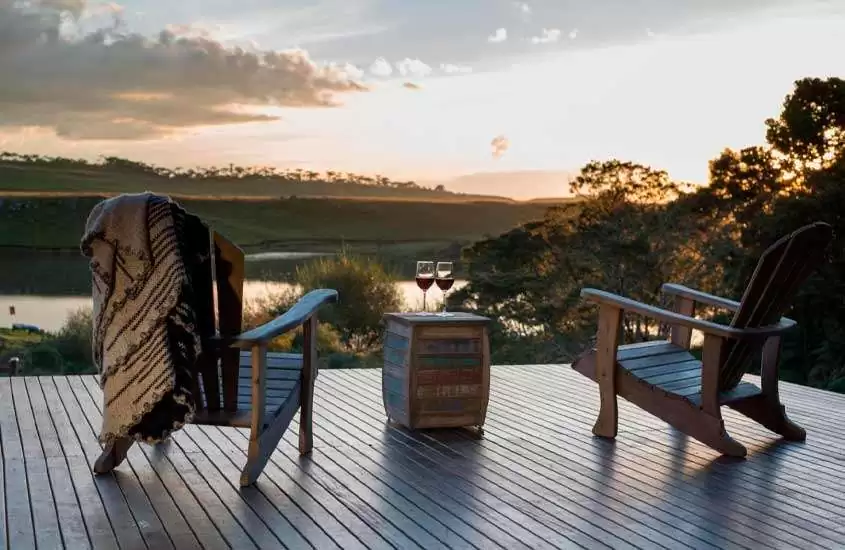 Durante o pôr do sol, deck com cadeiras, mesinha com taças de vinho, vista para o lago, árvores e área gramada