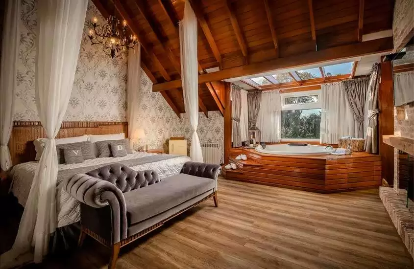 Quarto de hotel de luxo em Gramado com cama de dossel, lareira, lustre elegante, frigobar e banheira com deck de madeira
