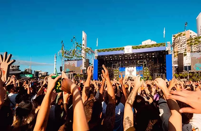 Em dia de sol, pessoas com as mãos para o alto se divertem em frente ao palco de um dos festivais de música brasil