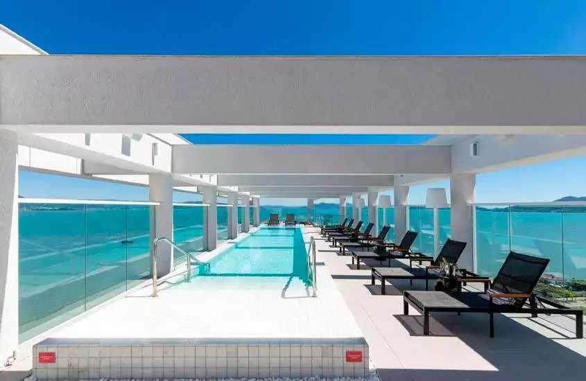 Em dia de sol, espreguiçadeiras ao redor de piscina em cobertura de hotel com vista para o mar