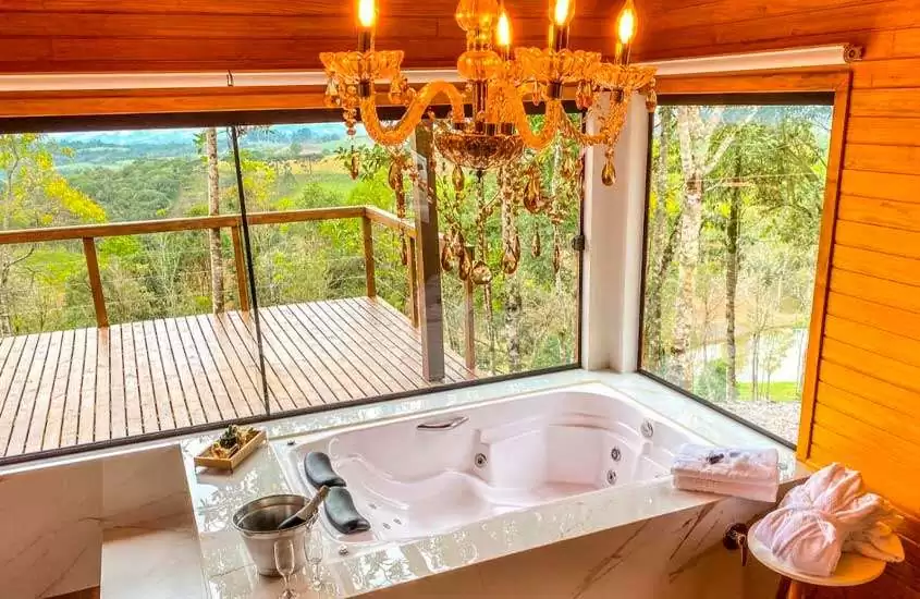 Em dia de sol, banheira de hidromassagem com toalhas, champagne, lustre e janelas grandes com vista do horizonte em chalé em santa catarina