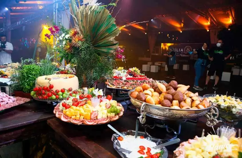 Mesa de ceia de ano novo em florianópolis com queijos, frutas, salgados e flores. Ao fundo, pessoas em pista de dança