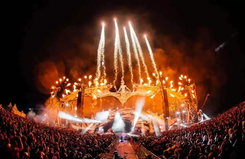 Durante a noite, multidão observa palco de um festivais de música brasil, iluminado por luzes amarelas. Ao fundo, show de fogos de artifício