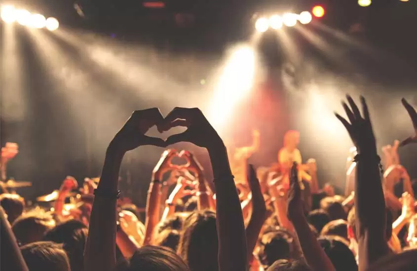 Durante show interno, pessoas levantando as mãos e fazendo corações para a banda se apresentando em palco iluminado por luzes coloridas