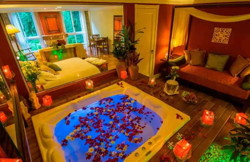 Quarto de um dos hotéis de luxo em gramado com banheira de hidromassagem, cama de casal e saleta com sofás, velas, quadros, vasos de planta, abajures e flores