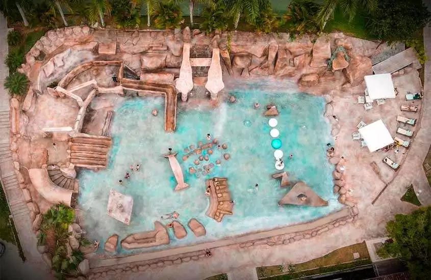 Durante o dia vista aérea de uma piscina com decorações em pedra, rodeada de espreguiçadeiras e guarda-sóis