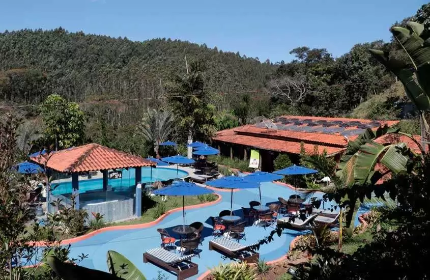Em dia de sol, vista aérea de piscina, espreguiçadeiras, mesas, cadeiras, jacuzzi e árvores em um dos hotéis fazenda do brasil