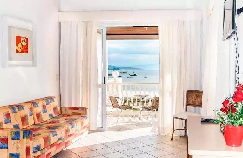 Em dia de sol, sala com sofá, cadeiras, mesas e janela grande pra varanda com vista para o mar, em um dos hotéis para passar o réveillon florianópolis