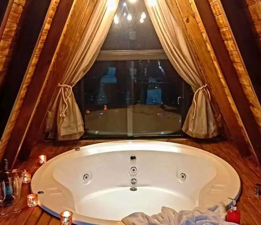 Durante a noite, banheira de hidromassagem com toalhas em deck de madeira, com taças, vinho em balde, velas, sabonete e janela grande acortinada