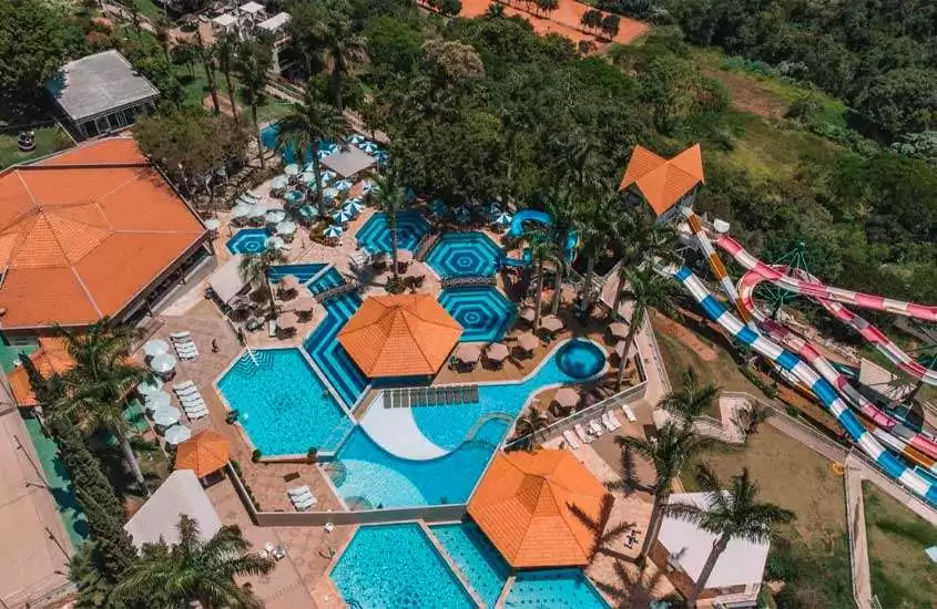 Em dia de sol, vista aérea de área de piscinas de hotel com bar molhado, toboáguas, árvores em volta, guarda-sóis, mesas e cadeiras.