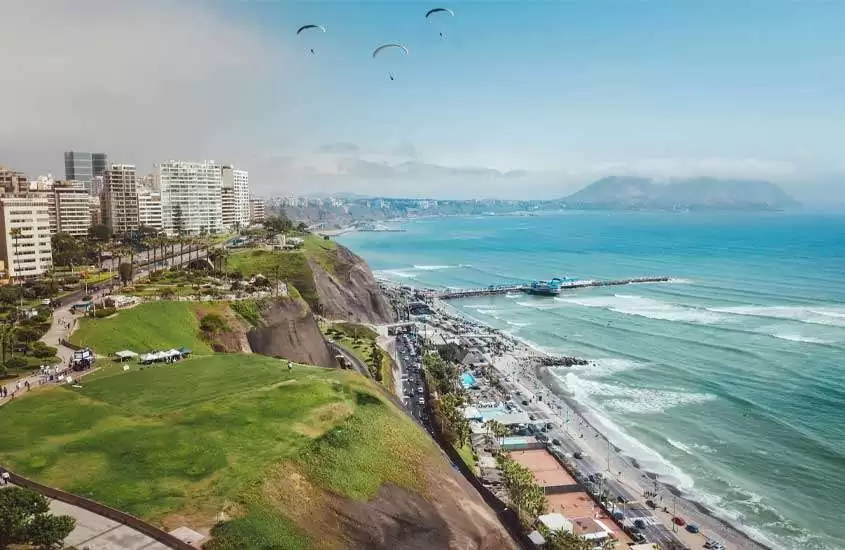 Durante o dia, vista panorâmica de prédios, casas, área gramada e pessoas em paraquedas às margens do mar em Miraflores, um bairro onde ficar em lima