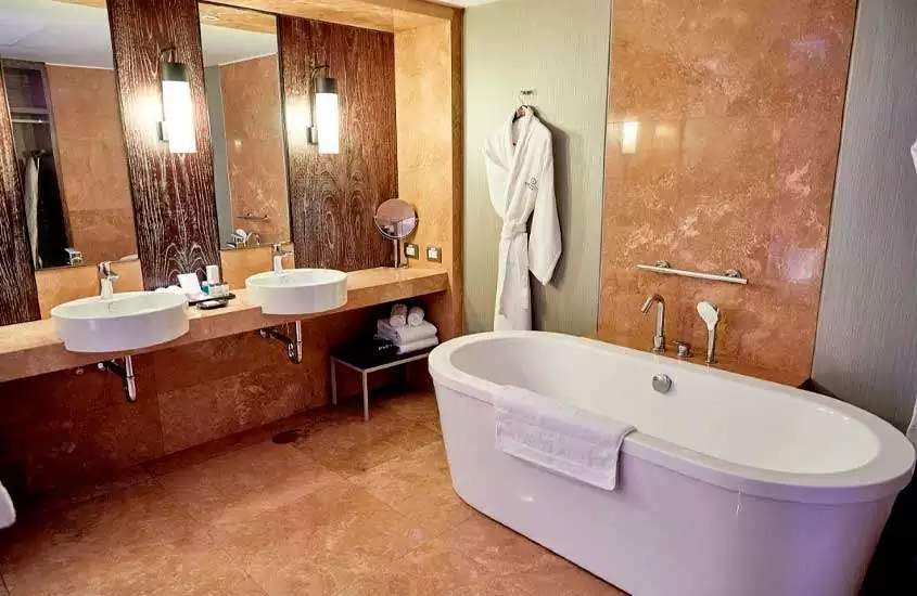 Banheiro de hotel com banheira, roupão, toalhas, duas pias, espelhos, luminárias e piso frio