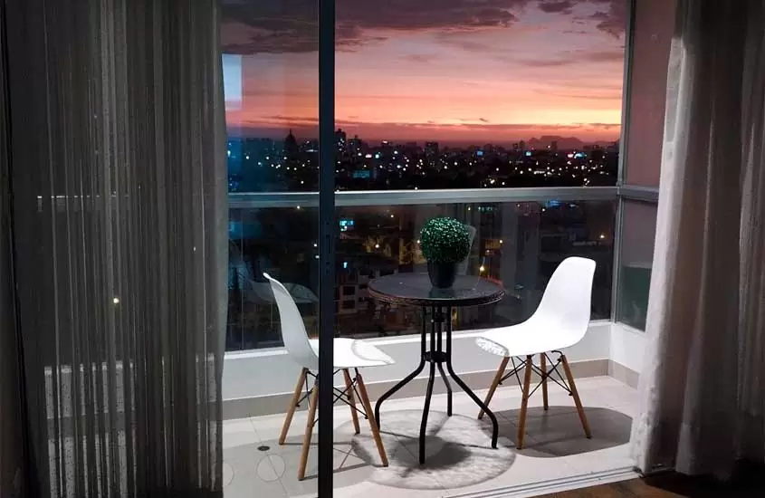 Durante o entardecer, mesa cadeiras e vaso de planta em varanda de suíte de hotel com vista da cidade