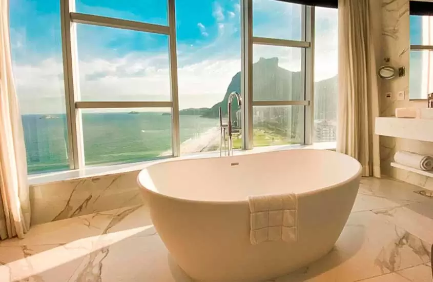 Durante o dia banheiro de um hotel para comemorar aniversário de casamento com banheira, toalhas, espelhos e janela grande com vista para o mar
