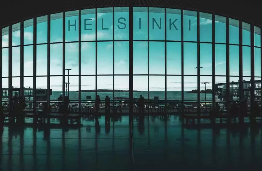 Em um dia de sol, interior de prédio em Helsinque com pessoas, mesas, cadeiras e mar atrás