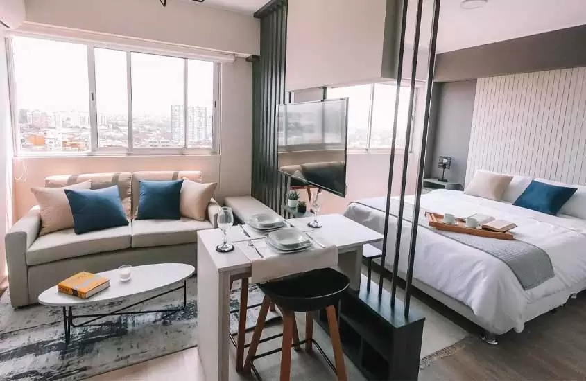 Em dia de sol, quarto de hotel em Lima com cama de casal, saleta de refeições, sofá mesa, TV e janelas grandes com vista da cidade
