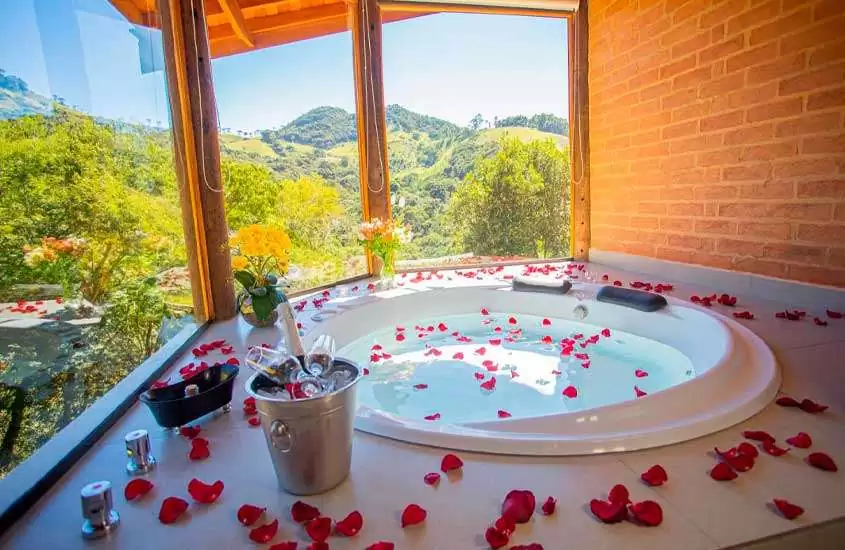 Em dia de sol, banheira de um hotel para comemorar aniversário de casamento com pétalas de rosa, balde de champagne com taças e janelas granes com vista do horizonte