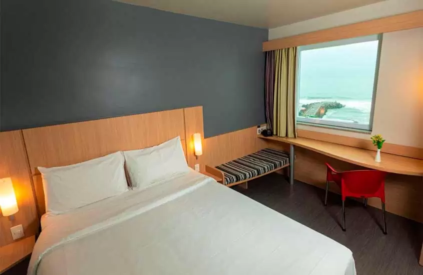 Quarto de um hotel na barra da tijuca beira-mar com cama de casal, cadeira, luminárias, banco, flor decorativa e janela acortinada