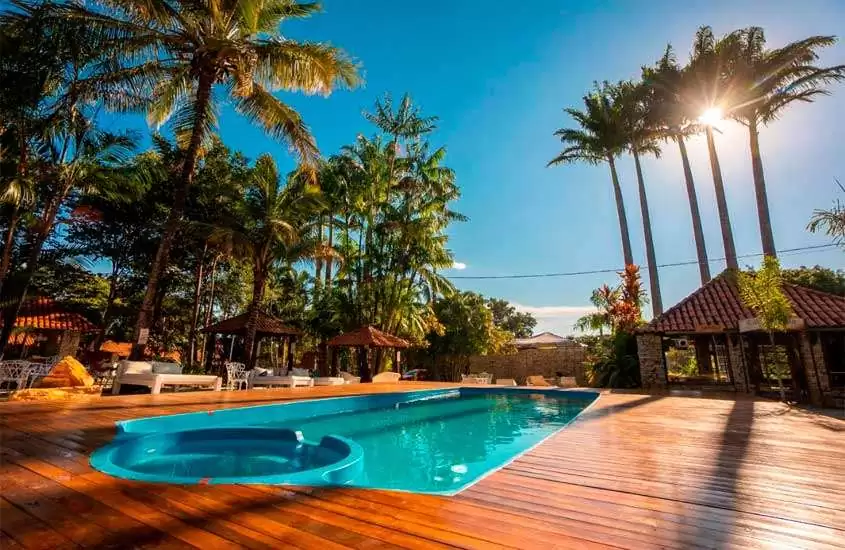 Em um dia de sol, área de lazer de um dos hotéis fazenda proximo a brasilia df com piscina, sofás, cadeiras, espreguiçadeiras, deck de madeira, coqueiros e hotel ao redor