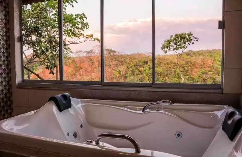 Em uma tarde nublada, banheiro de um dos hotéis fazenda proximo a brasilia df com banheira, janela grande e árvores na frente