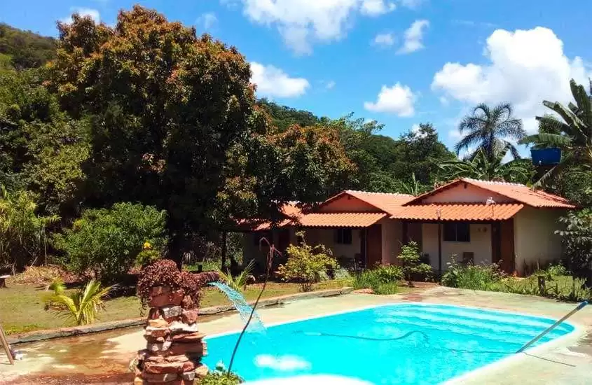 Em um dia de sol, área de lazer de um dos melhores hotéis fazenda proximo a brasilia com piscina, cascata, árvores, flores e hotel do lado