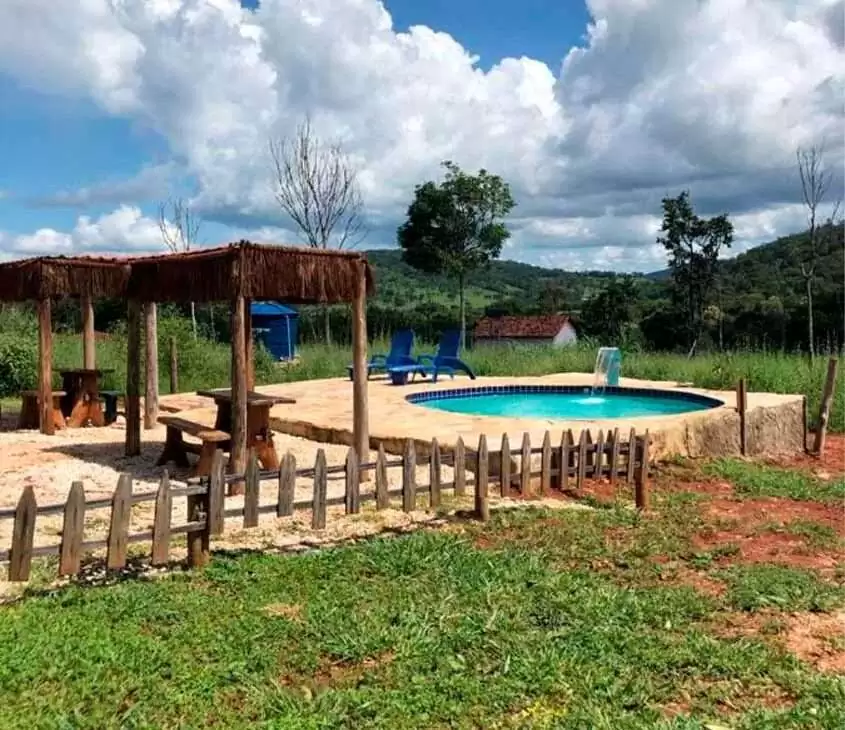 Em um dia de sol, área de lazer de um hotel fazenda perto de brasília com piscina, cascata, bancos, mesas, espreguiçadeiras, parte gramada e árvores ao redor