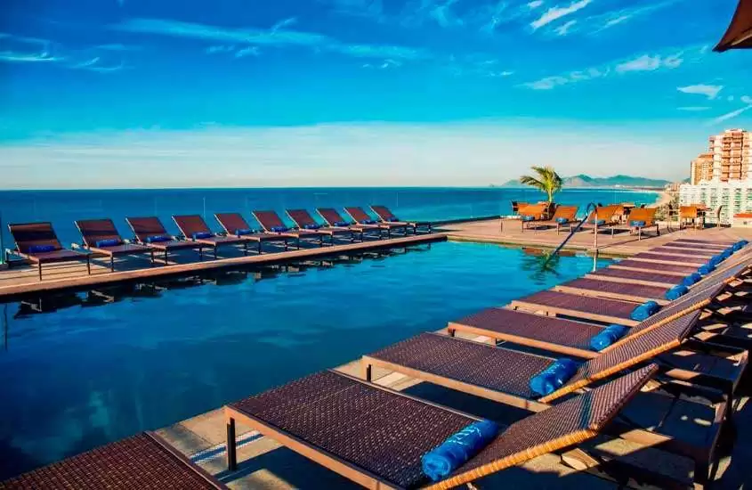 Em um dia de sol, área de lazer de um dos hotéis na Barra da Tijuca em frente à praia com espreguiçadeiras, toalhas azuis e piscina
