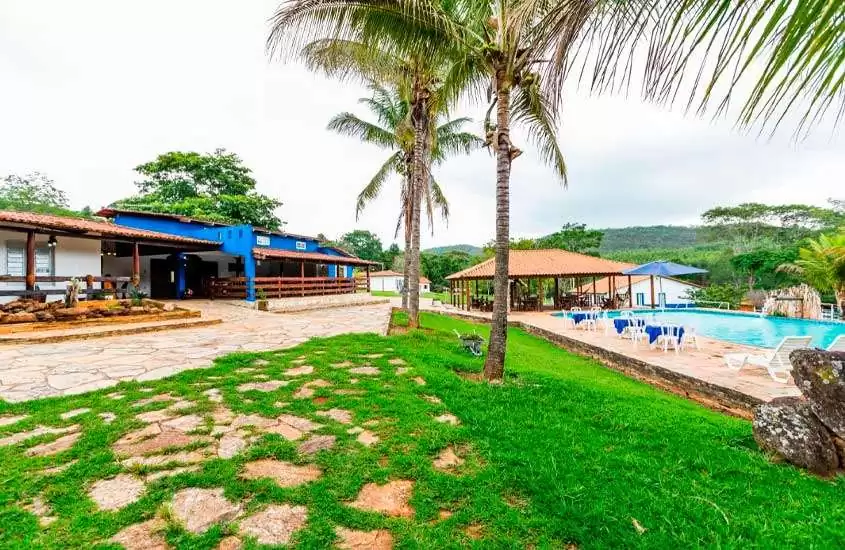 Em um dia nublado, área de lazer de hotel fazenda perto de brasília com piscina, mesas, cadeiras, espreguiçadeiras, parte gramada, árvores e hotel ao redor