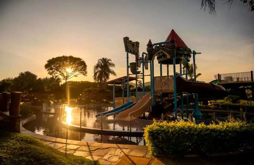 Durante o pôr do sol, área de lazer de um hotel fazenda perto de brasília com parquinho, piscina, parte gramada, árvores e arbustos